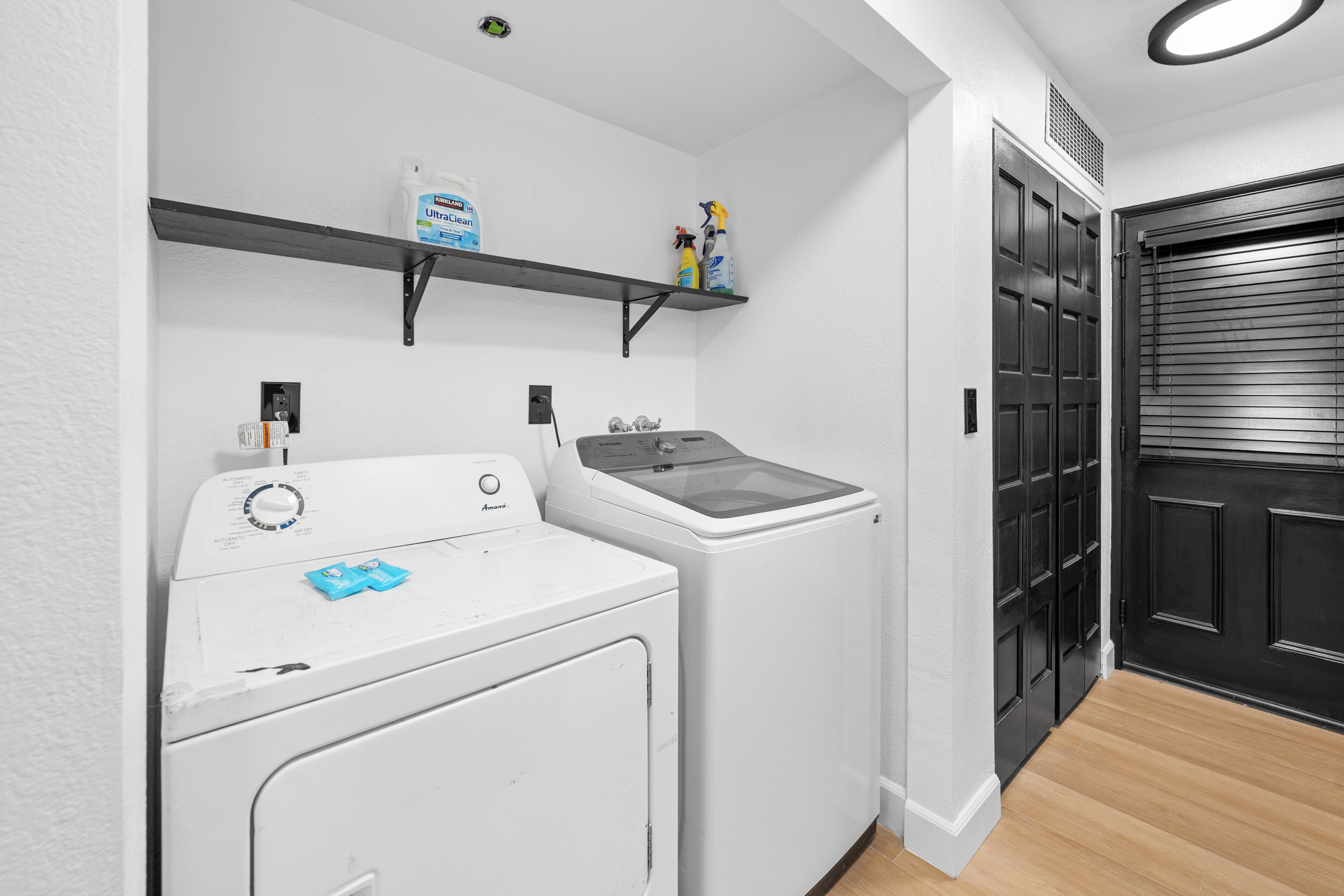 A laundry area with a front-loading washer and dryer, a shelf with cleaning supplies, and a black door.