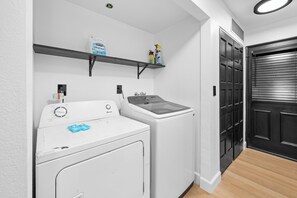 A laundry area with a front-loading washer and dryer, a shelf with cleaning supplies, and a black door. - Summit Mountain Cabin (Big Bear Lake)
