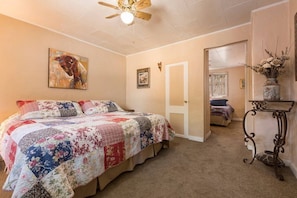 A bedroom with a large bed, a ceiling fan, a door leading to another room, and a small table with a vase.