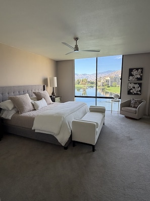 A bedroom with a large bed, a chair, a small table, and a view of a lake and mountains. - Spacious Contemporary -- Striking Mountain Views 2B/2BA in Coveted Desert Island (Rancho Mirage)
