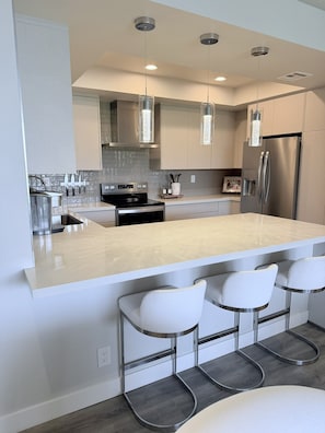 Une cuisine moderne dotée d’un grand îlot central, d’appareils électroménagers en acier inoxydable, de luminaires suspendus et de tabourets de bar.