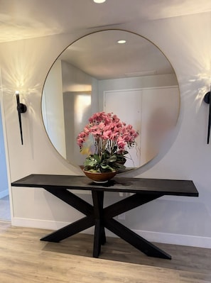 Une pièce avec un grand miroir rond posé sur un socle en bois foncé, une plante en pot aux fleurs roses et, au fond, une porte blanche.