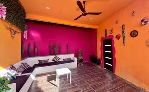 Une pièce avec un mur d’un orange vif, un ventilateur de plafond, un banc blanc agrémenté de coussins et une petite table blanche.