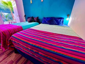 A room with two beds, each with a colorful striped bedspread, a blue wall, and a small window.