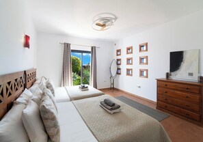 Ein Schlafzimmer mit zwei Betten, einer Holzkommode, einem Deckenventilator und Blick auf Grünfläche.