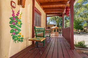 Eine Holzterrasse mit einer Bank und einem Tisch, eine kleine Holzhütte und eine Mauer mit einer Uhr und gemalten Blumen.