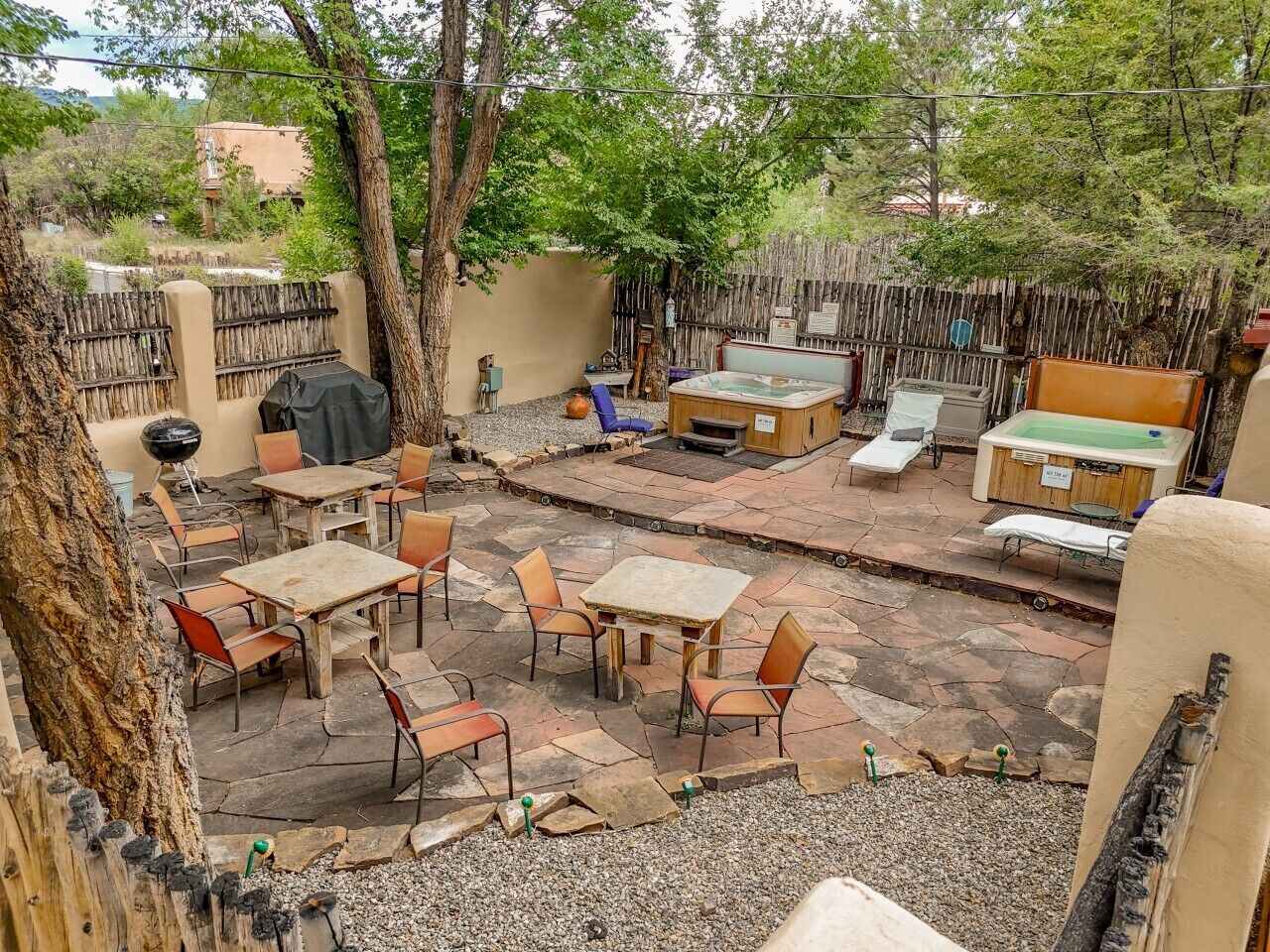 An outdoor patio with stone flooring, wooden tables, and chairs, surrounded by trees and a wooden fence.