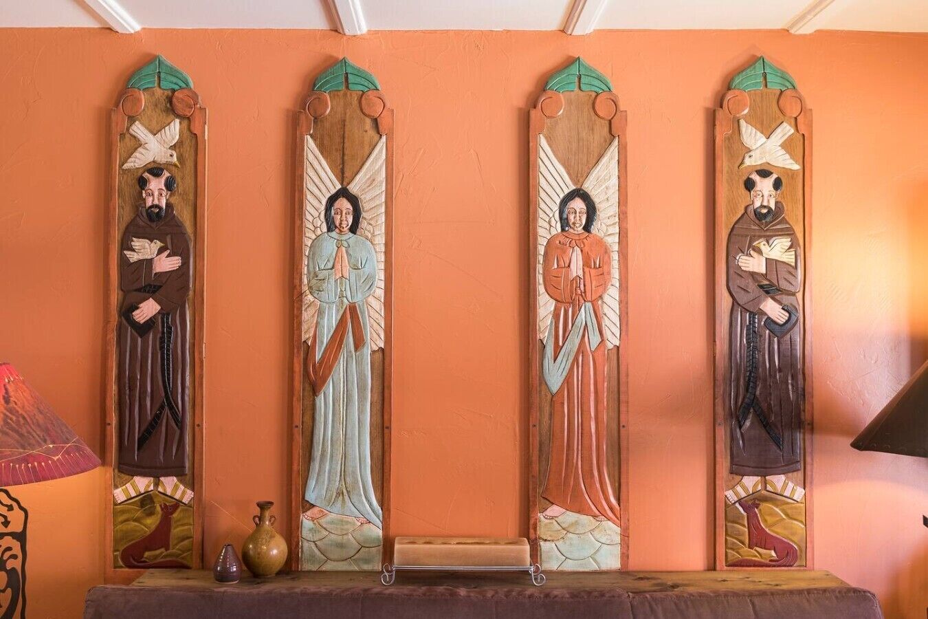 Un mur composé de quatre panneaux verticaux, chacun représentant une figure ailée tenant une colombe, et un vase placé à gauche.