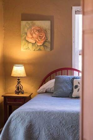 A bedroom with a bed, a nightstand with a lamp, a wall-mounted painting of a rose, and a window.