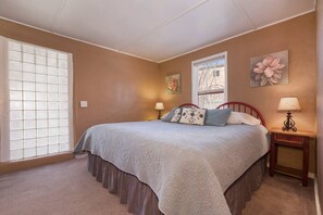 Un dormitorio con una cama, una ventana con persianas, una mesita de noche con lámpara y un cuadro de flores en la pared.