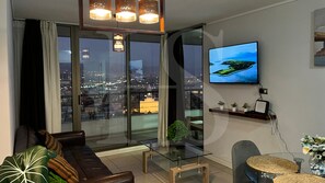 A modern living room with a large window, a flat-screen TV, a sofa, a glass coffee table, and a small plant.
