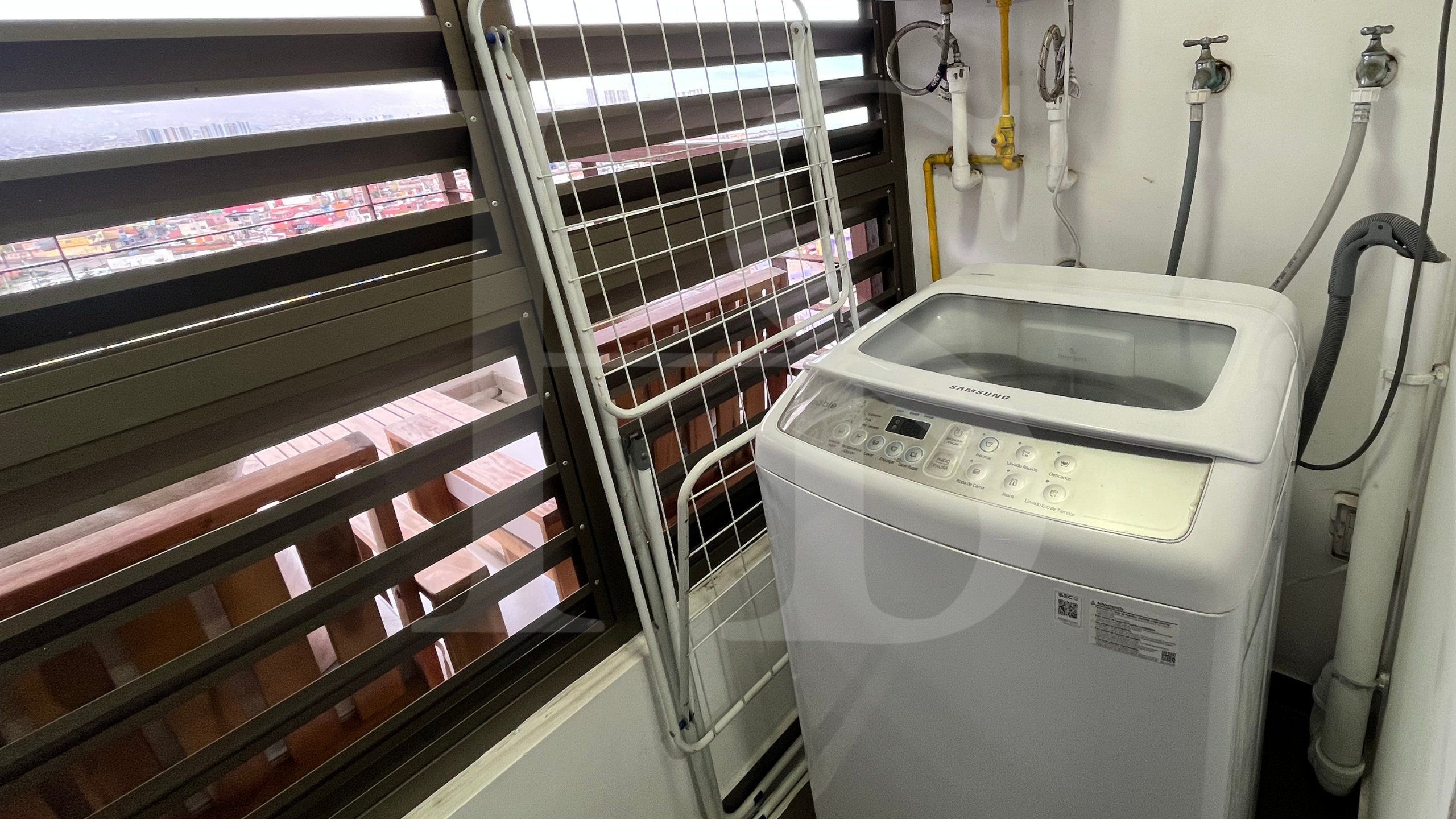 Un lavadero con una lavadora Samsung, un tendedero con ropa doblada y una ventana con persianas.