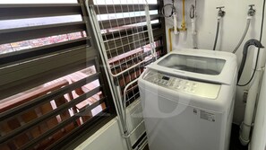 Un lavadero con una lavadora Samsung, un tendedero con ropa doblada y una ventana con persianas.