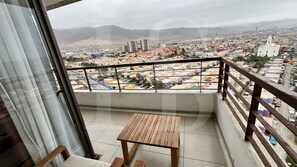 A balcony with a wooden table and chairs, overlooking a cityscape with buildings and mountains.