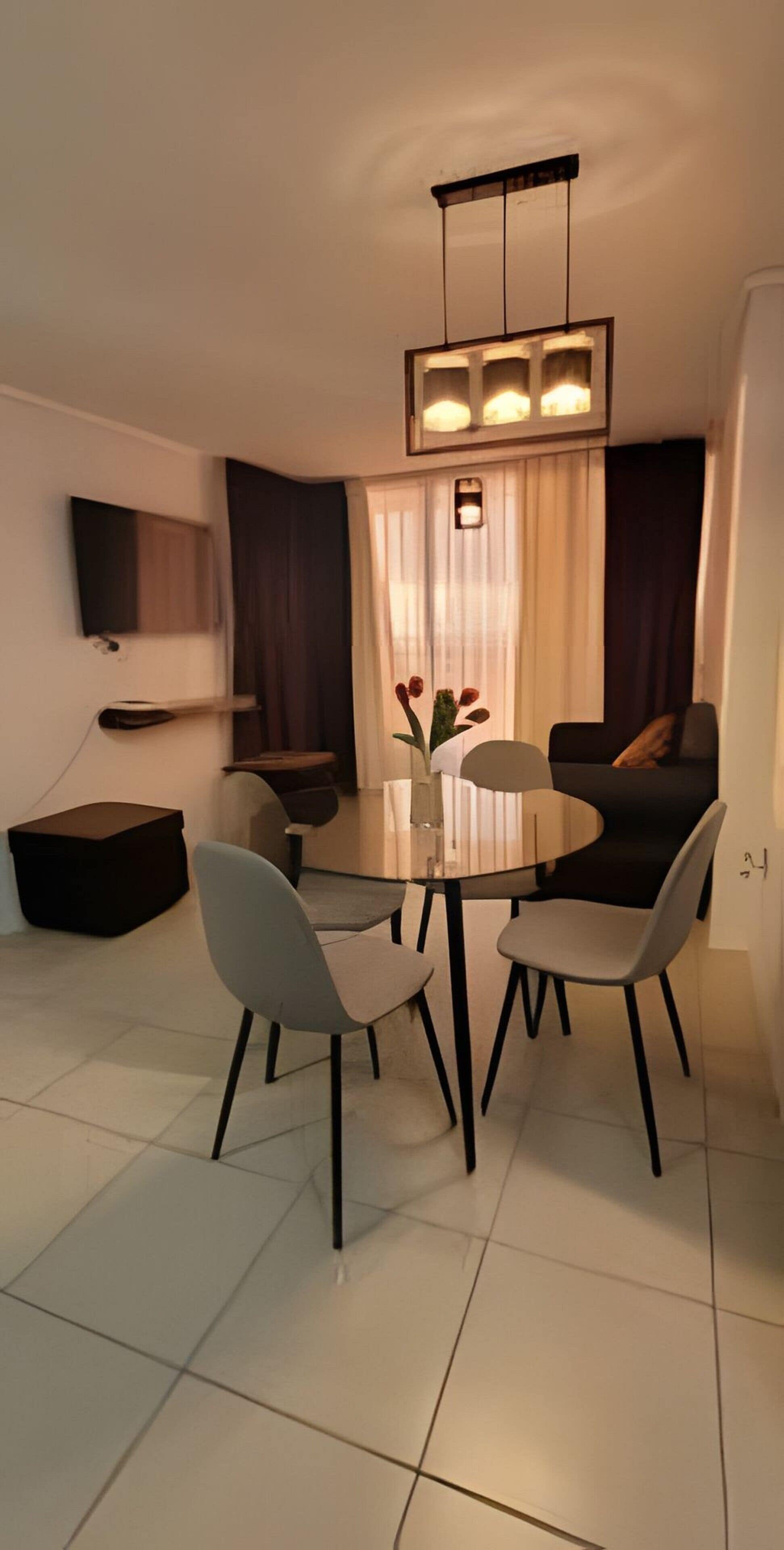 Un comedor moderno con una mesa redonda, cuatro sillas, un florero con flores, un televisor de pantalla plana en la pared y un sofá al fondo.