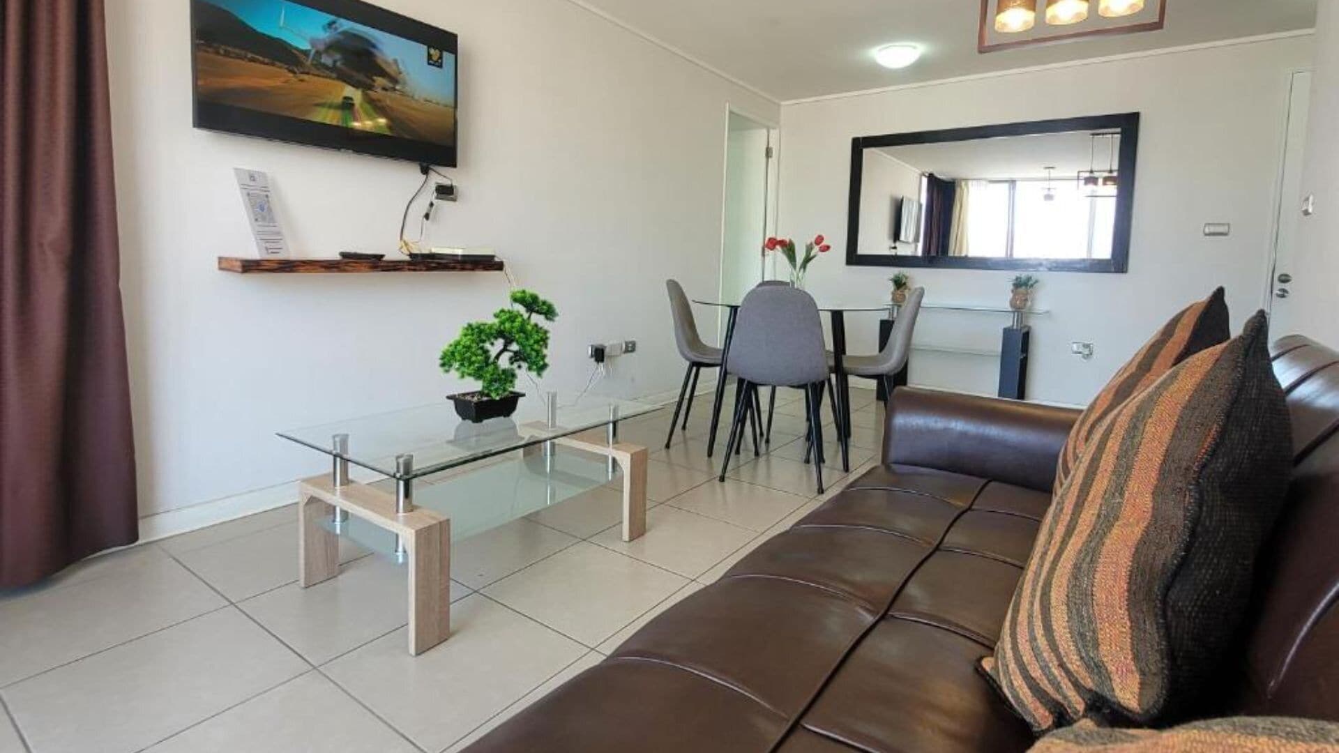 Una sala moderna con una mesa de centro de vidrio, un sofá de cuero marrón y una zona de comedor con sillas.