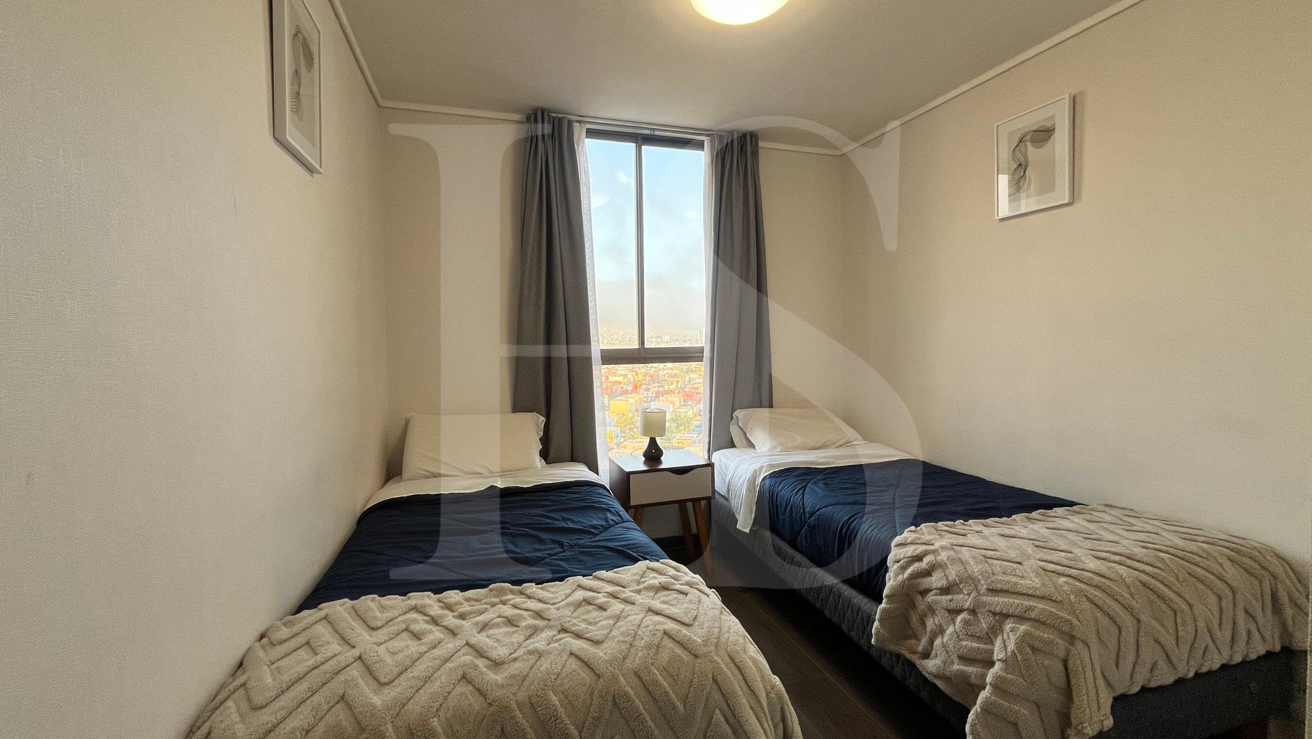 A small, narrow room with two beds, a nightstand, a window with curtains, and a wall-mounted light fixture.