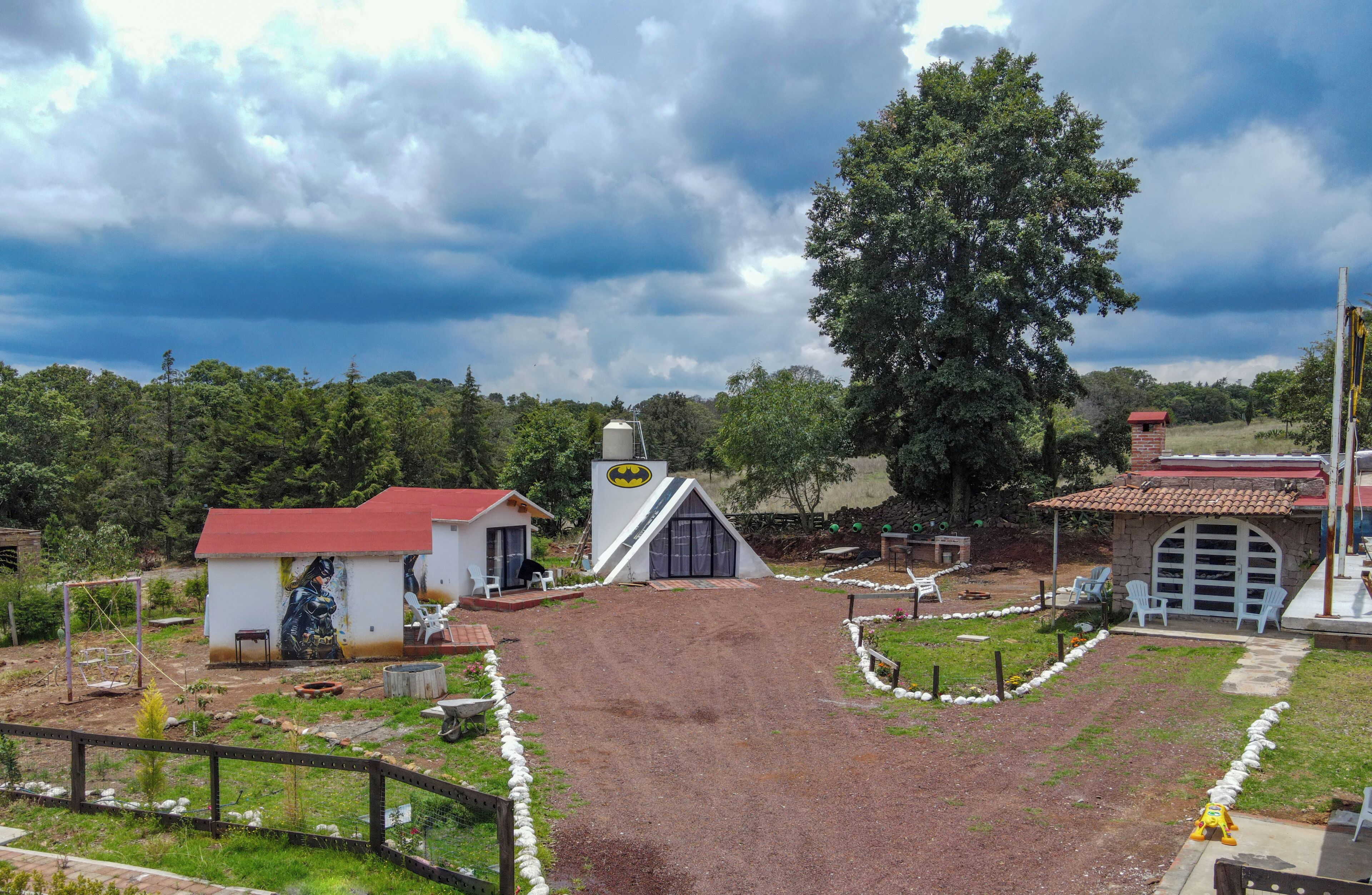 Una propiedad rural con camino de tierra, un pequeño edificio blanco con techo rojo y un árbol grande.
