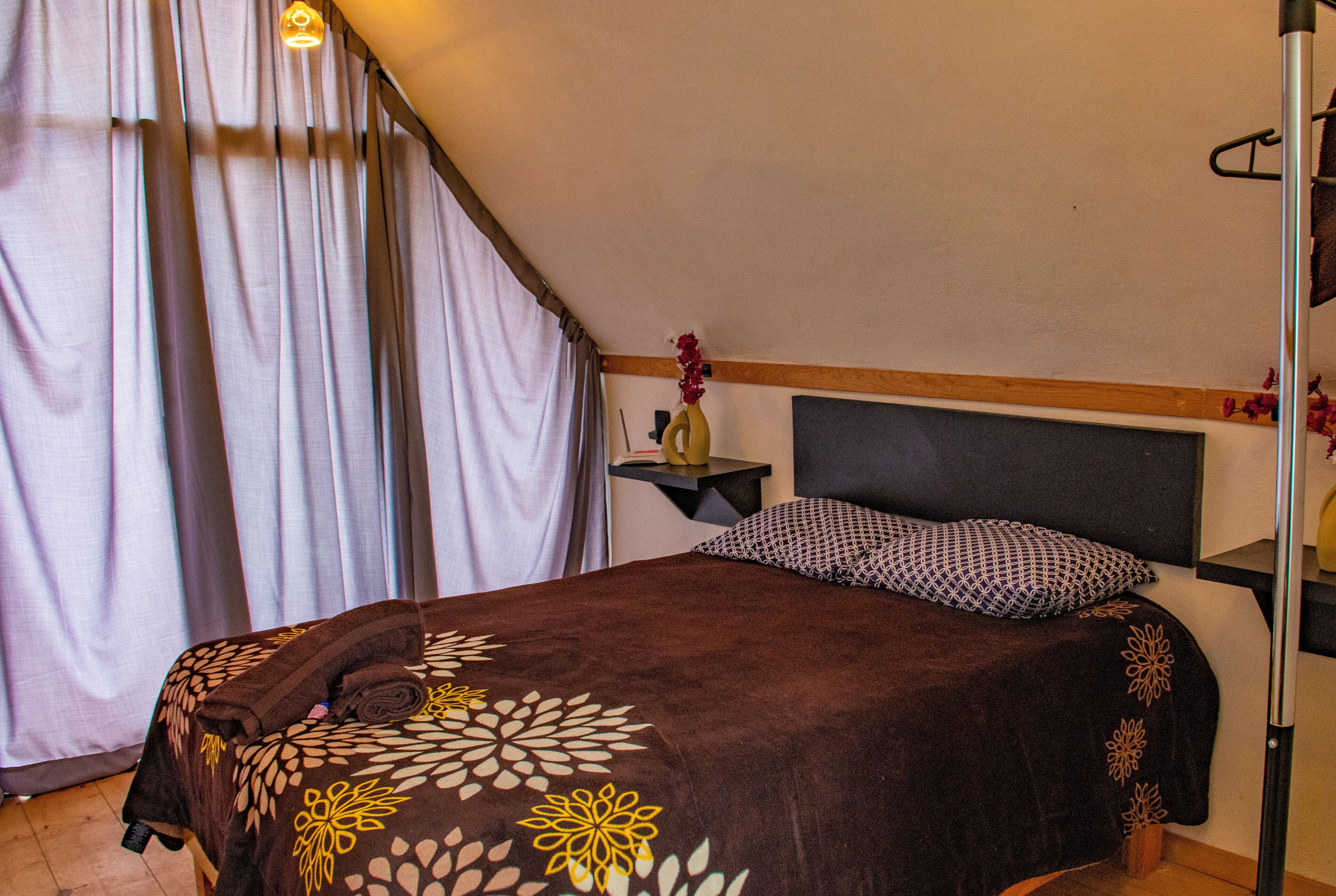 A bedroom with a bed, a nightstand, a wall-mounted shelf, and a window with curtains.