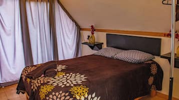 A bedroom with a bed, a nightstand, a wall-mounted shelf, and a window with curtains.