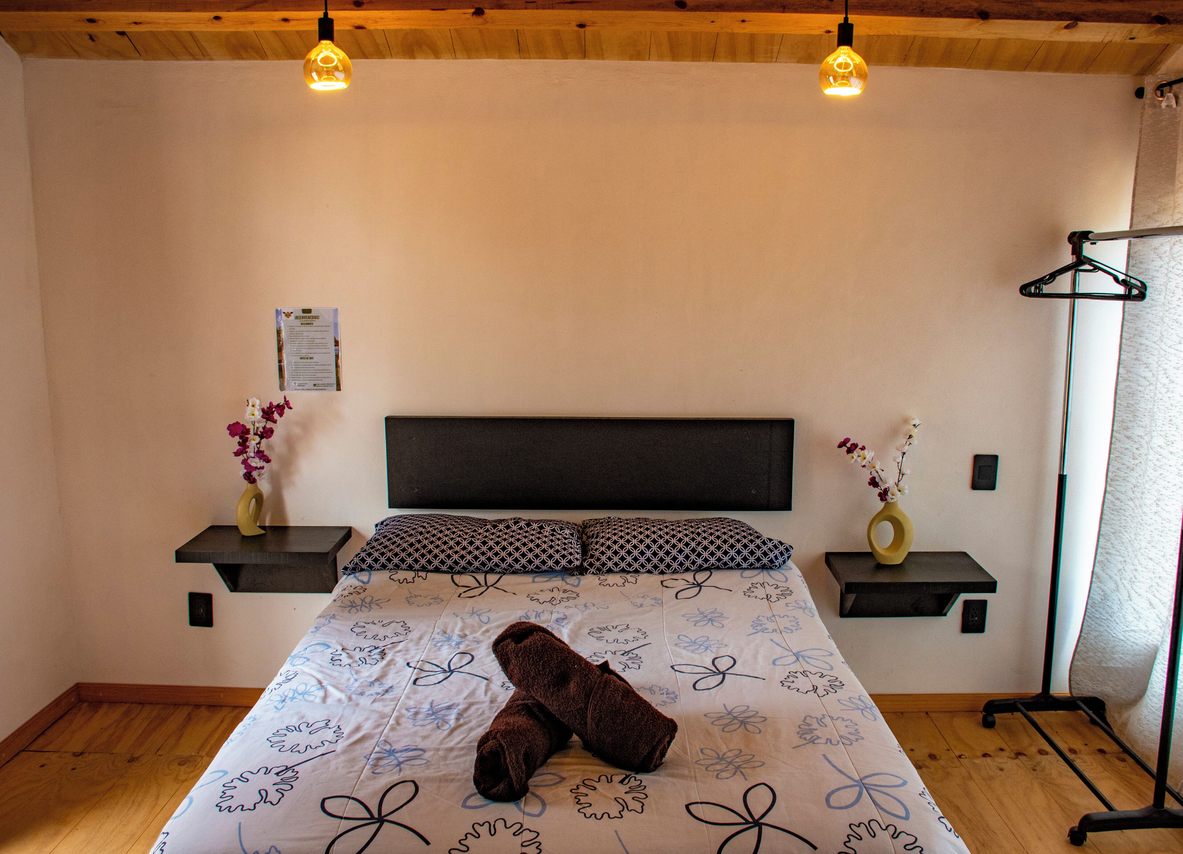 Una habitación de hotel con una cama, dos mesitas de noche, una lámpara de pared y un techo de madera.