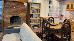 A cozy living area with a brick fireplace, a wooden bookshelf, a dining table with chairs, and a kitchen area with a window.