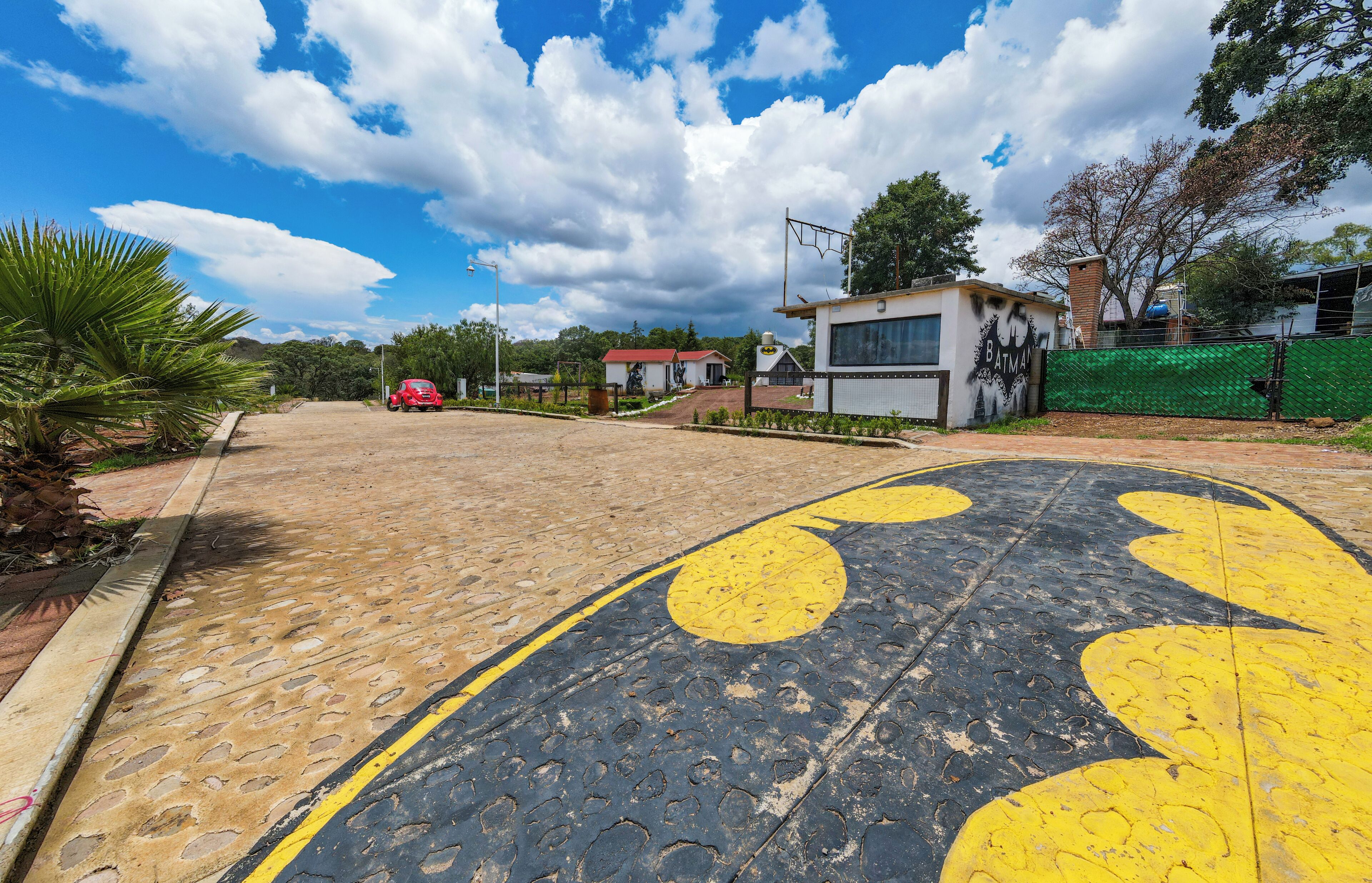 Una carretera pavimentada con marcas amarillas y negras, un auto rojo estacionado al lado, y un edificio con el logo de Batman.