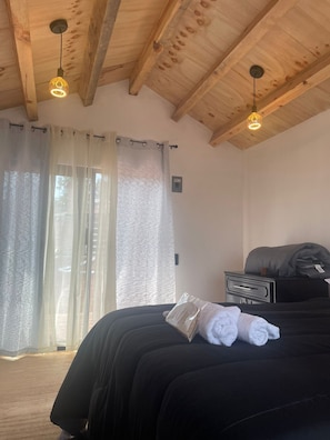 Une chambre avec un plafond en bois, un lit avec une literie noire, une table de chevet et une fenêtre ornée de rideaux légers.
