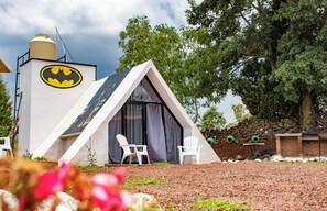 Un edificio triangular con el logo de Batman, dos sillas blancas afuera y un muro de piedra al fondo.