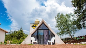 A triangular building with a large window and a Batman logo, two white chairs outside, and a gravel ground.