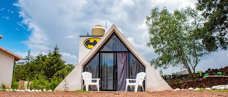A triangular building with a large window and a Batman logo, two white chairs outside, and a gravel ground.
