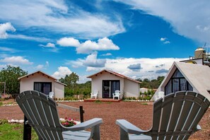 Two small, single-story buildings with a clear sky and clouds above. There are two white chairs in the foreground, and a garden area with rocks and plants.