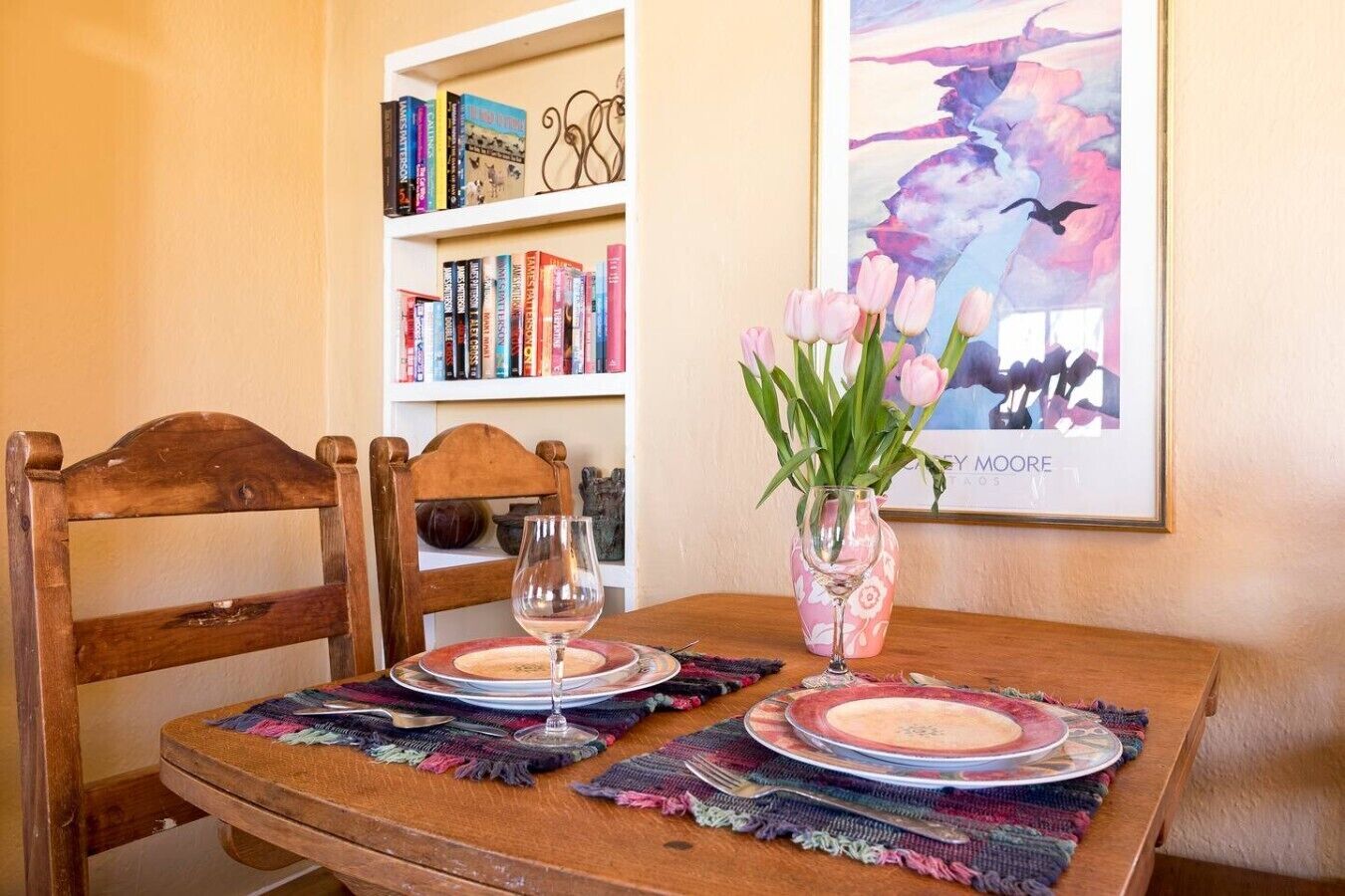 Une table à manger avec un vase de fleurs, un verre à vin et une assiette. On aperçoit une bibliothèque remplie de livres et un tableau encadré accroché au mur.
