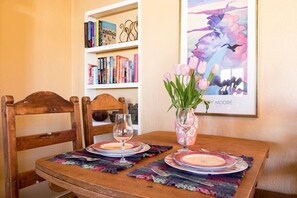 A dining table with a vase of flowers, a wine glass, and a plate. There is a bookshelf with books and a framed picture on the wall.