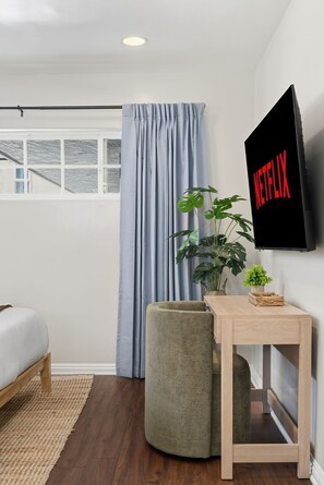 A modern room with a bed, a small table, a potted plant, and a wall-mounted TV displaying 'NETFLIX'. - Newport Beach & Bay Oasis 2 King Beds, Smart TVs, AC Bike Rentals & Beach Gear (Newport Beach)