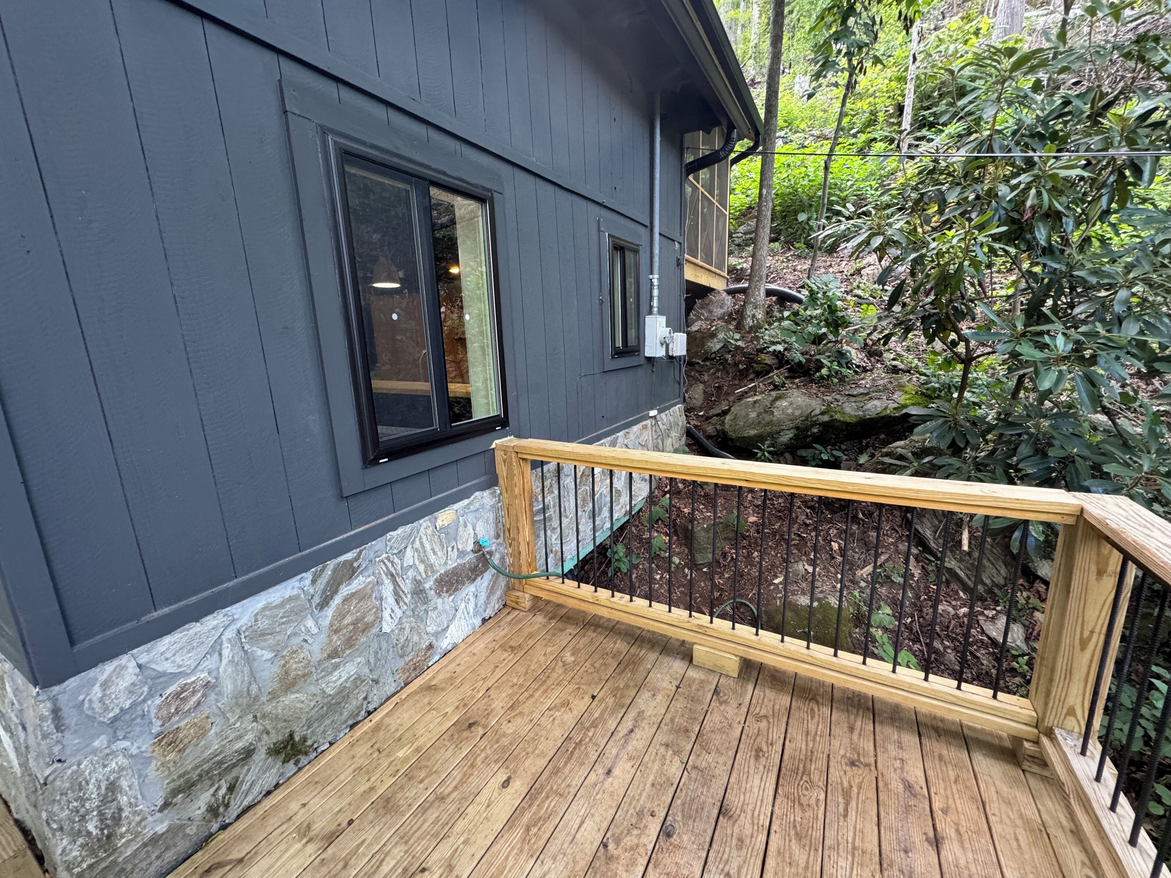 Une terrasse en bois avec une fondation en pierre, une fenêtre à cadre noir et une rambarde en métal.