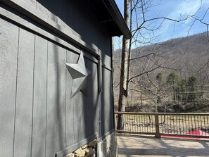 Ein hölzernes Gebäude mit sternförmiger Verzierung, einem Steinfundament und Blick auf eine bewaldete Landschaft, in der Ferne ein Gebäude mit rotem Dach sichtbar.