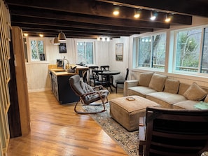 Un interior acogedor con piso de madera, un sofá, un sillón mecedor, una mesa de comedor y una zona de cocina con ventanal.