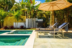 Ein Schwimmbecken mit Liegestühlen und Grillplatz, umgeben von tropischen Pflanzen.