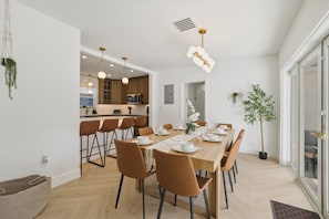 A modern dining area with a long table set for six, surrounded by chairs, with a kitchen in the background. - Fox Eden Resort 8Bd Pool Spa Oasis (Fort Lauderdale)