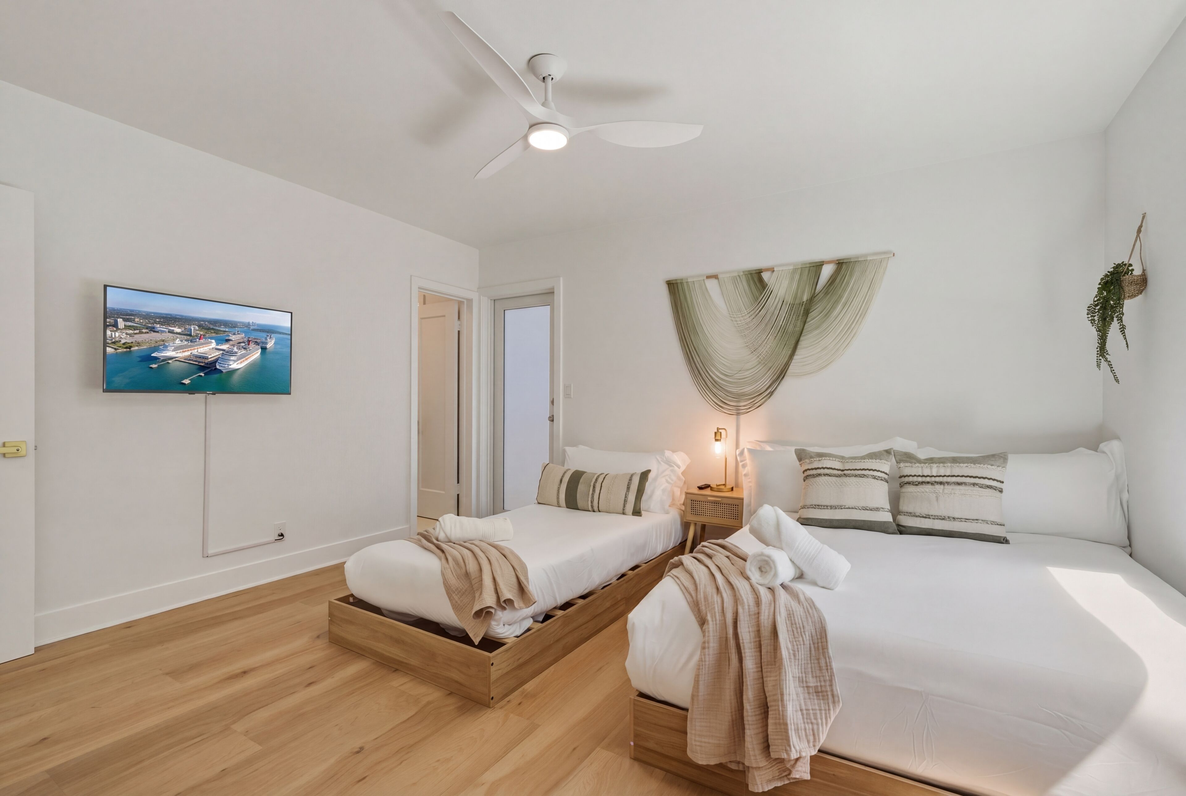 A modern hotel room with a bed, a wooden bench, a ceiling fan, a wall-mounted TV, and a view of a harbor.