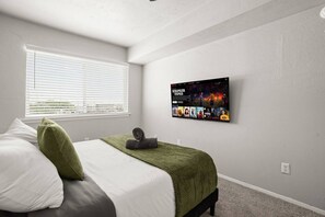 Ein Schlafzimmer mit einem Bett, einem Fenster mit Jalousien, einem an der Wand befestigten Fernseher, der einen Film zeigt, und einer Steckdose an der Wand.