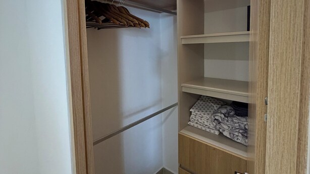 Ein kleiner, abgeschlossener Schrank mit Holztüren und Regalen, der Kleidung zum Aufhängen und gefaltete Kleidungsstücke enthält.