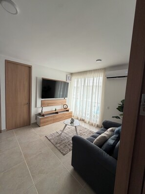 Una sala de estar moderna con una televisión de pantalla plana, muebles de madera, un sofá y un amplio ventanal con cortinas.