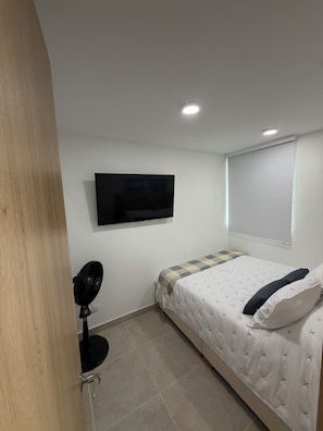 A bedroom with a bed, a wall-mounted TV, a ceiling fan, and a window with blinds.