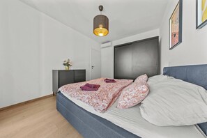 A bedroom with a bed, a bedside table with a vase of flowers, a wardrobe, and a hanging light fixture. - Eurovea Tower, Danube View, Balcony & Parking (Ružinov)