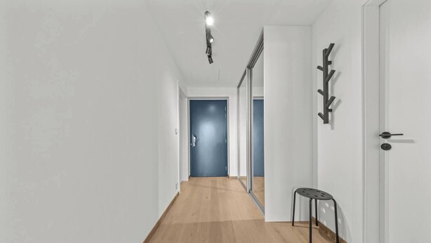 Un passadís amb sòl de fusta, una cadira, un penjallabecs i una porta que dona a una altra habitació.