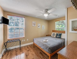 A bedroom with a bed, a ceiling fan, a TV, and a window with blinds.