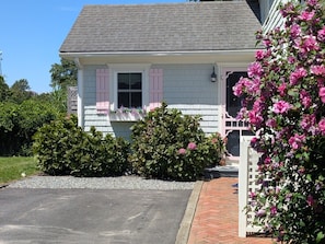 A two-story house with a sloped roof, a front porch, and a small garden area with shrubs and flowers. - Quaint property right in town (New Shoreham)