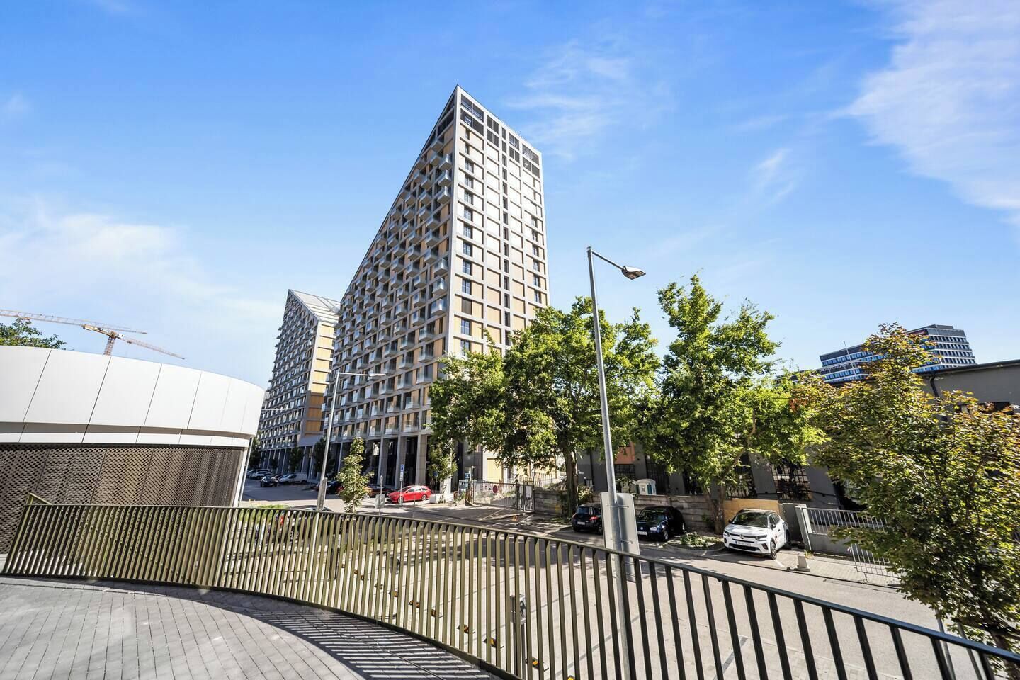 A modern urban building with a prominent corner design, a pedestrian walkway, and a street with parked cars.